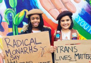 Two Radical Monarchs hold signs in front of a mural painting during a protest march