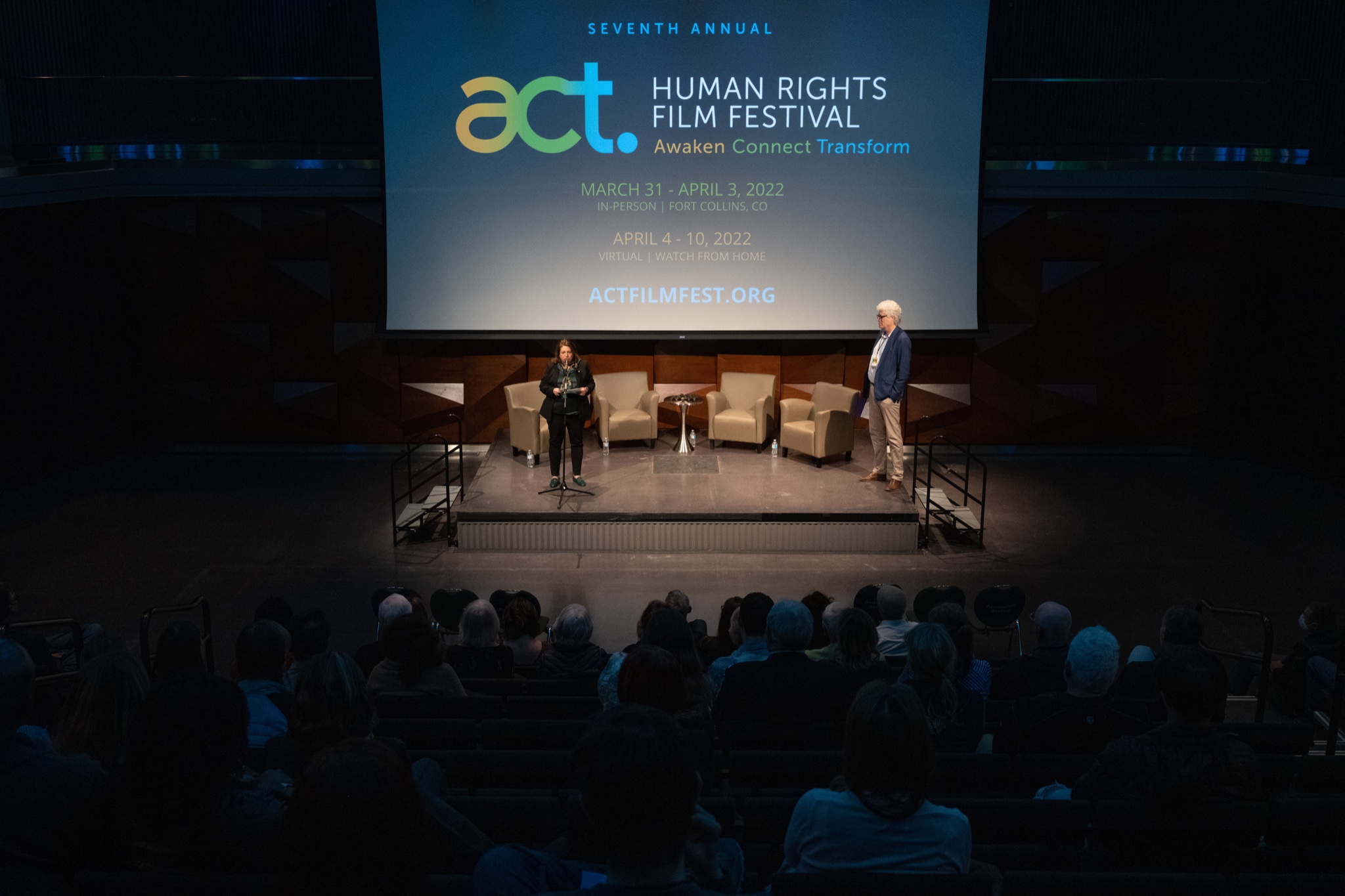 A man and woman stand on stage in a darkened theater with "Seventh Annual ACT Human Rights Film Festival" displayed on the screen behind them