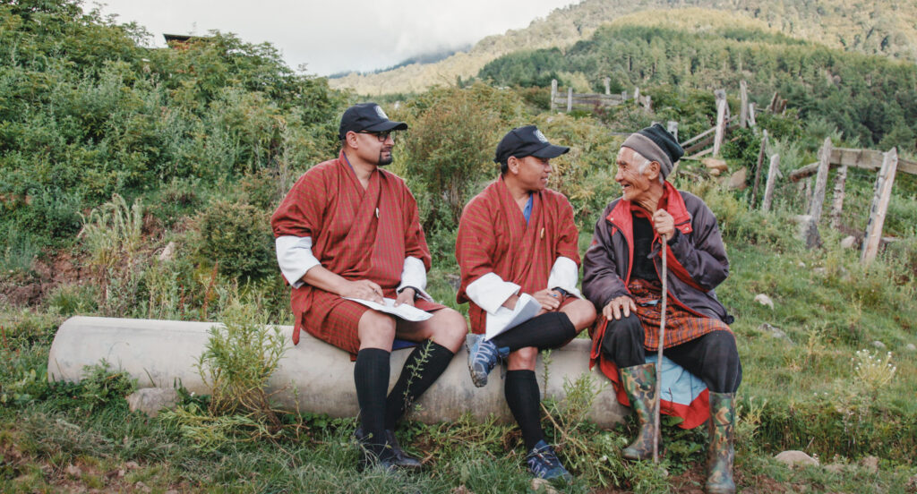 Three Bhutanese men sit on a log in the countryside. Mountains in the background. 