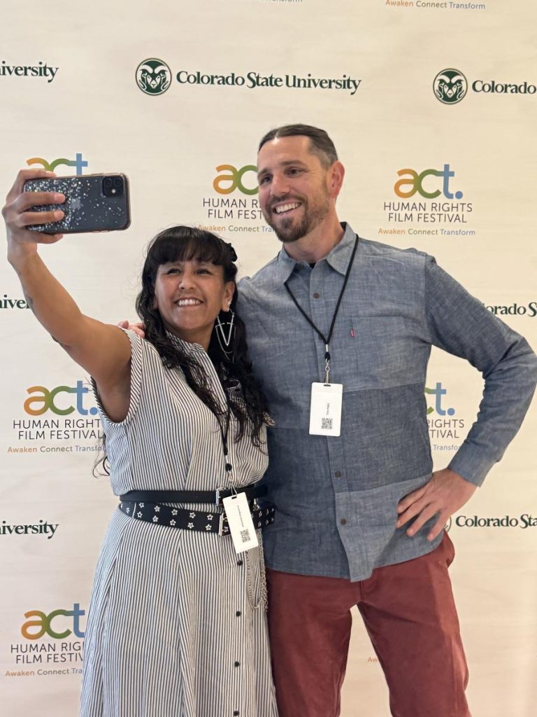 A woman and a man stand together in front of the ACT Human Rights Film Festival back drop and take a selfie together. 